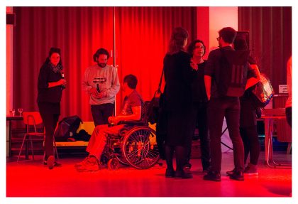Bildbeschreibung: In einem Raum mit einem rot leuchtenden Licht, sieben Personen in zwei Gruppen miteinander chatten mit Getränken in ihren Händen. Einige Leute stehen, während ein anderer im Rollstuhl sitzt.