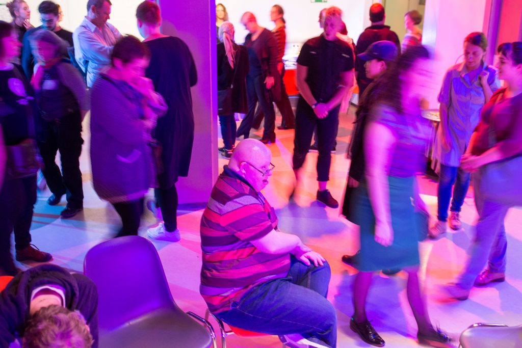 A room full of people moving around, one person in the middle sitting still on a chair, purple light in the room