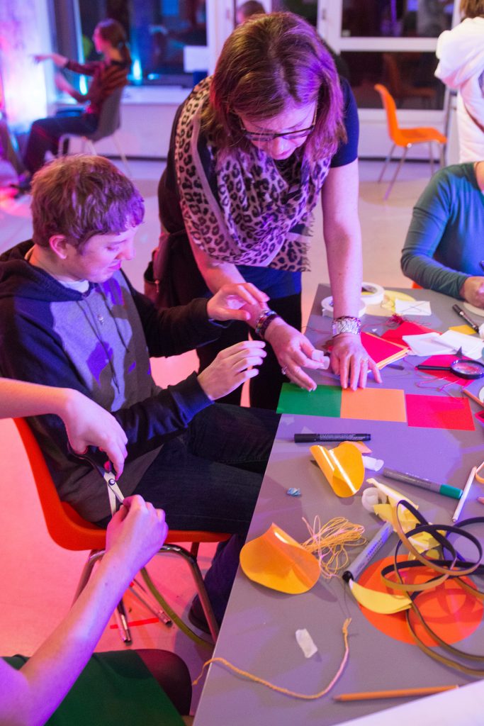People sitting and standing around table, together creating using different crafts materials that lay on the table