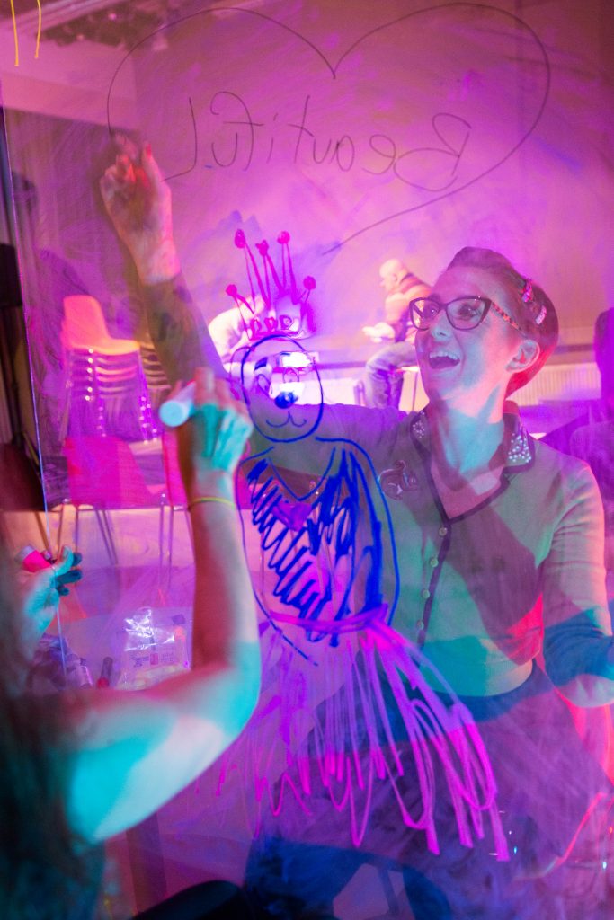Two persons drawing simultaneously on a transparent board on opposite side of each other, one is drawing a character with a crown, the other one is smiling, soft purple light in the room