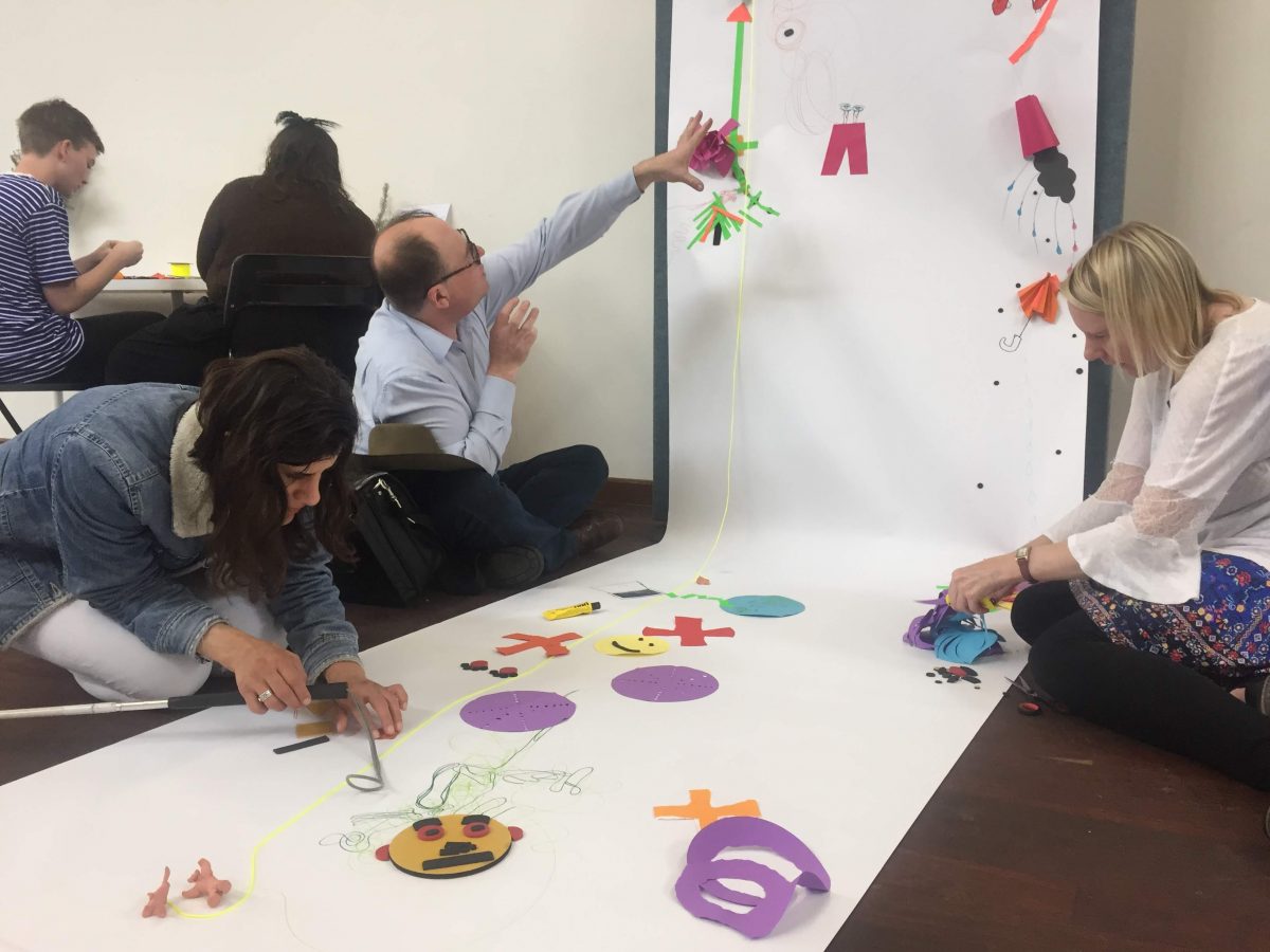 One man and two women attaching on a white paper roll colorful objects made of different materials