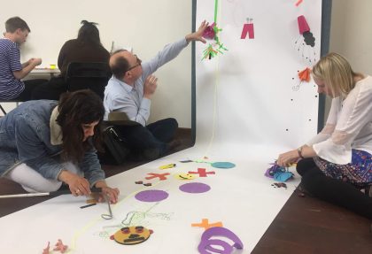 One man and two women attaching on a white paper roll colorful objects made of different materials