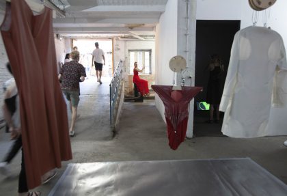 Blick in Ausstellung: Im Vordergrund hängen Kleider von der Decke. Im Hintergrund Menschen auf einer Rampe und Performerin im roten Kleid auf Sockel sitzend.