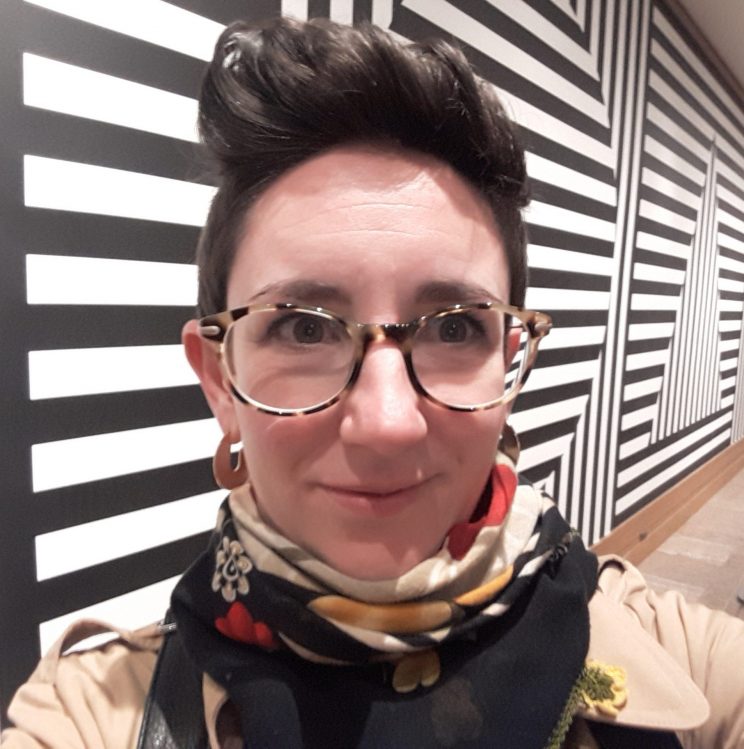 Ein Porträt von Kate Brehme, einer weißen Frau mit kurzen, dunkelbraunen Haaren, Brille und einem bunten Schal, steht vor einem schwarzweißen, geometrischen Wandgemälde von Sol Le Witt.