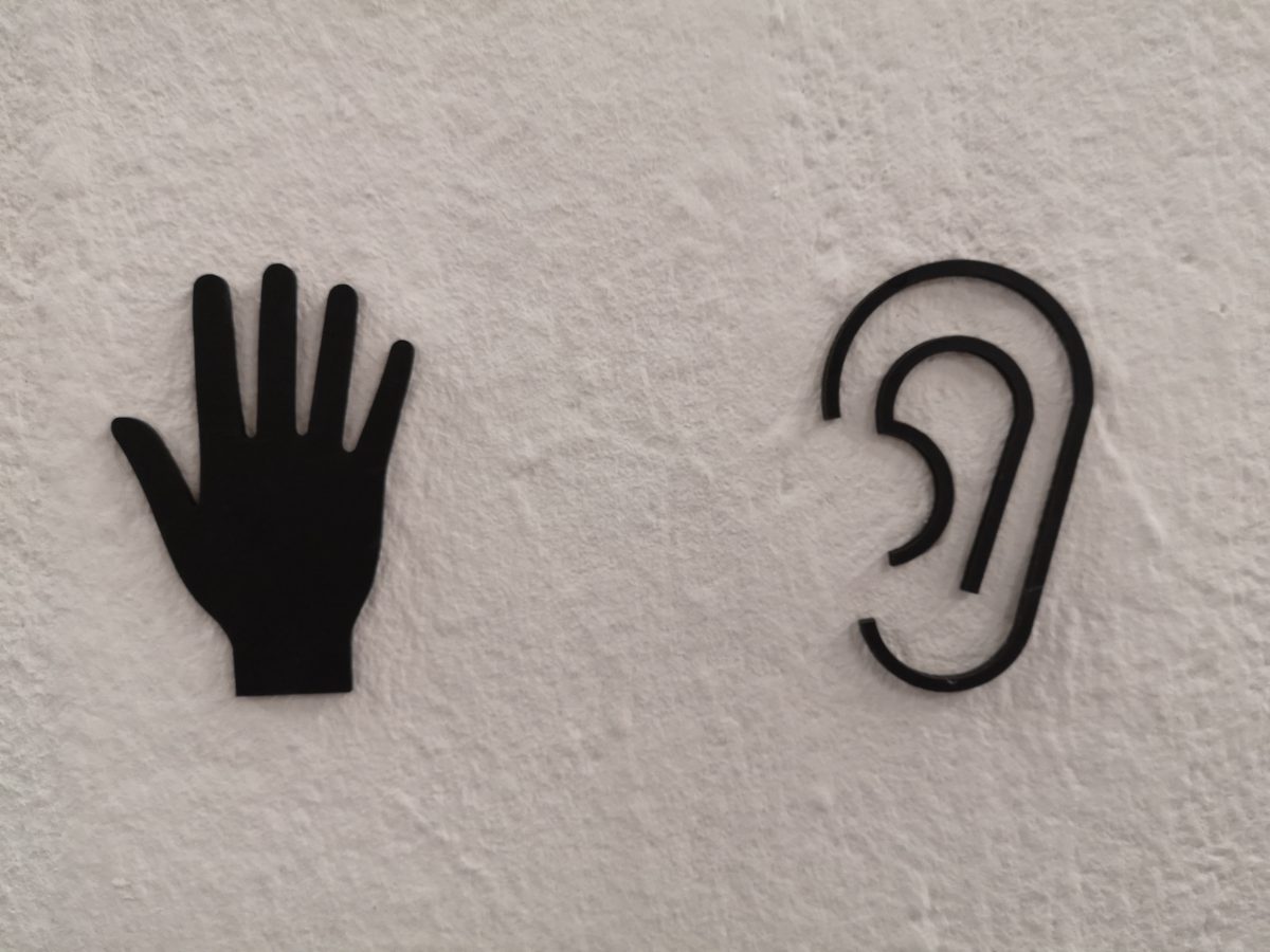 An image of two black tactile signs on a white wall. On the left is a hand with fingers splayed, On the right is an outline of an ear.