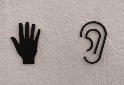 An image of two black tactile signs on a white wall. On the left is a hand with fingers splayed, On the right is an outline of an ear.