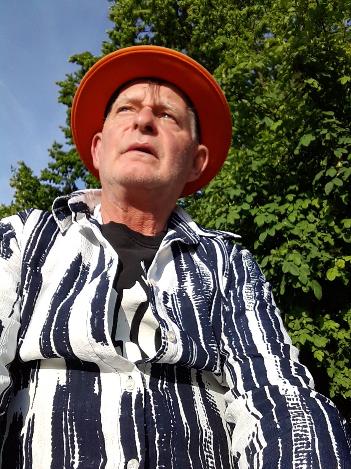 Macks wird von unten fotografiert, wir schauen zu ihm hoch, während er an uns vorbeischaut. Er ist ein weißer Mann in den 60ern, der einen leuchtend orangefarbenen Hut und ein weiß-schwarz gestreiftes Hemd trägt.