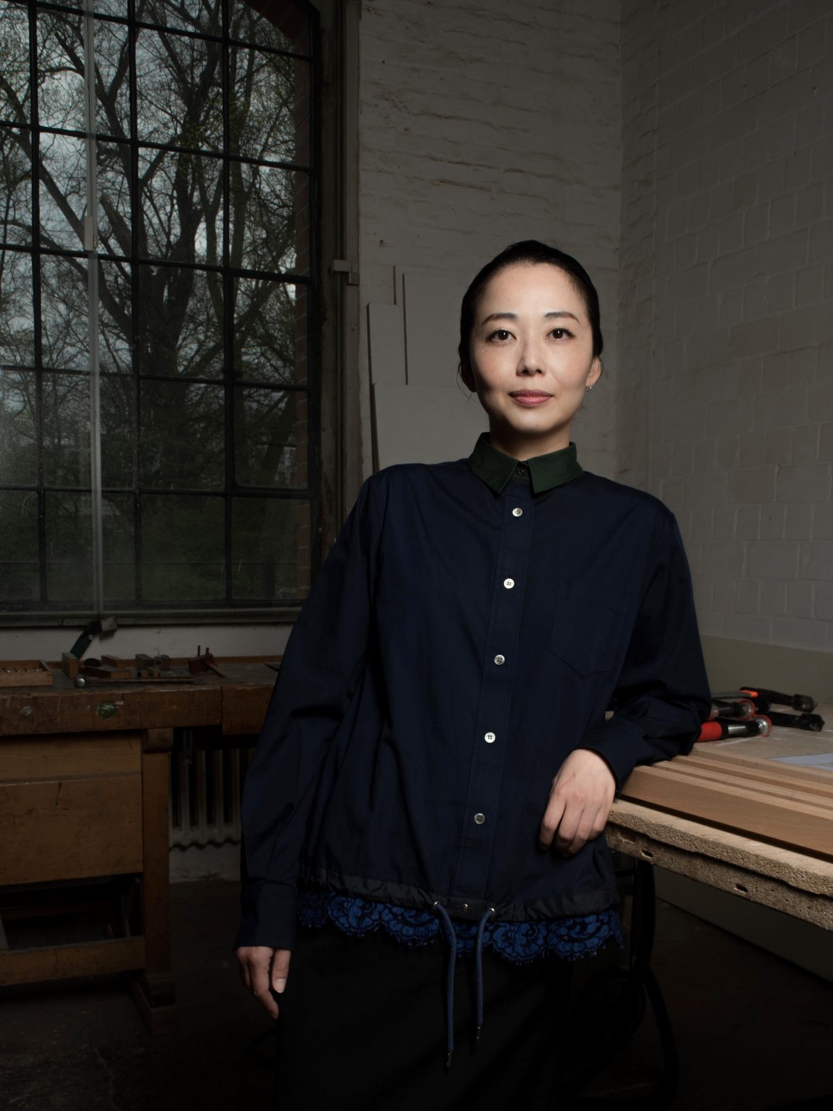 Asako, eine zierliche Japanerin, starrt kühn in die Kamera, während sie steht und ihren linken Arm auf einen Tisch stützt.