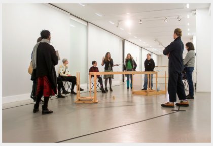 A group of people sitting and standing in a gallery around an installation made of wood, are listening to someone giving a talk. Eine Gruppe von Menschen, die in einer Galerie um eine Installation aus Holz herum sitzen und stehen, hört jemandem zu, der einen Vortrag hält.