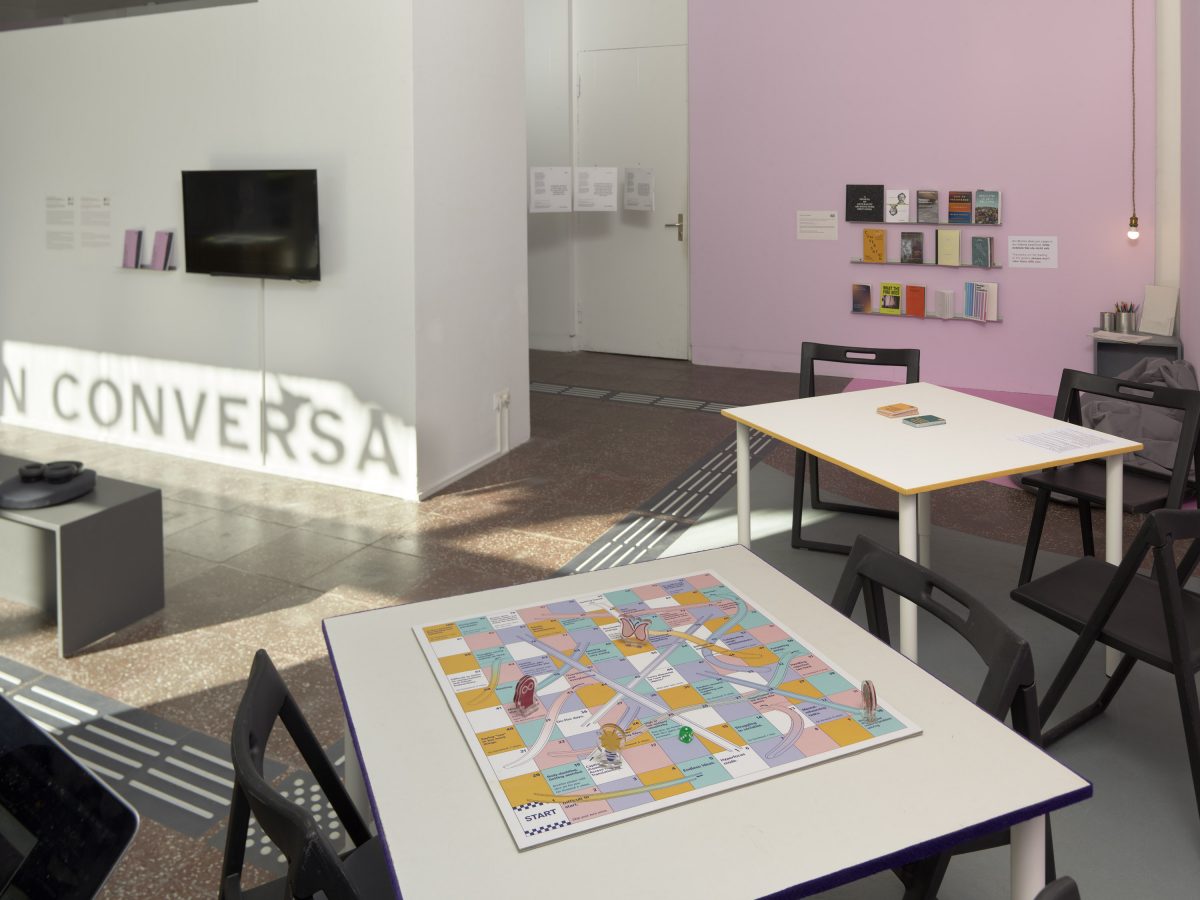 A white and pink gallery space with a tactile floor system, a video, a bookshelf wiht books and two tables with a board game and card game.