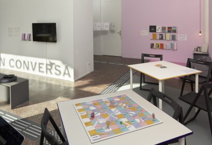 A white and pink gallery space with a tactile floor system, a video, a bookshelf wiht books and two tables with a board game and card game.